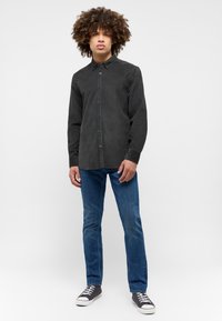 Camicetta in velluto a coste nera con bottoni sul davanti, maniche lunghe e colletto, abbinata a jeans slim-fit blu e sneakers nere con suola bianca.