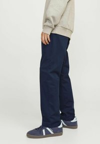 Person iført marineblå afslappede bukser, grå sweatshirt og marineblå sneakers med hvide striber, stående på en lys baggrund.