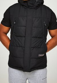 Vadderad svart väst med dragkedja fram och fickor, utrustad med huva och quiltad yta. Logopatch på den nedre framsidan.