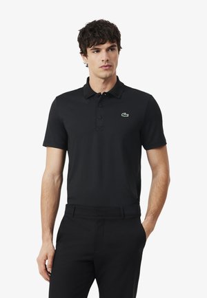 Uomo che indossa una polo nera a maniche corte con un piccolo logo di coccodrillo verde sul petto a sinistra, abbinata a pantaloni neri.