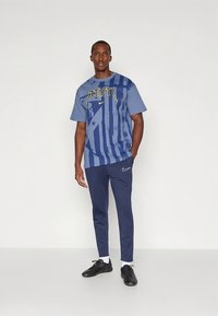 Zila grafiskā T-krekls ar rakstainām svītrām un logotipu, kombinēts ar tumši ziliem sporta biksēm un melnām sporta kurpēm. Gluda auduma un ikdienišķa piegriezuma.