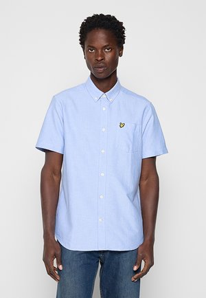 Mann trägt ein hellblaues, kurzärmliges Hemd mit Knopfleiste und einem gelben Vogel-Logo auf der Brusttasche, kombiniert mit blauen Jeans.