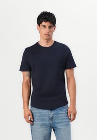 Marineblaue Baumwoll-T-Shirt mit kurzen Ärmeln, Rundhalsausschnitt und dezentem Logo-Detail. Kombiniert mit hellen Jeans aus Denim, durchgehend glatte Textur.