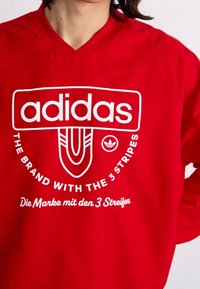 Red sweatshirt made of cotton blend featuring a prominent white Adidas logo and text. Simple crew neck design with ribbed details.