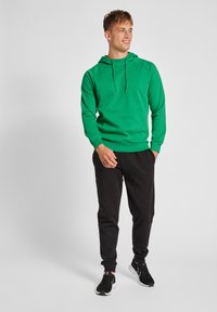 Grüne Kapuzenjacke mit Kordelzug und gerippten Bündchen, kombiniert mit schwarzen Jogginghosen und schwarzen Turnschuhen. Schlichtes Design, weiches Material und lockere Passform.