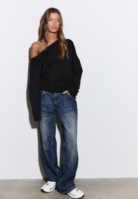 Črni ohlapni pulover z enim izpovednim ramenom, kombiniran s svetlo modrimi, posivljenimi kavbojkami s pravimi hlačnicami in belimi atletskimi supergami.