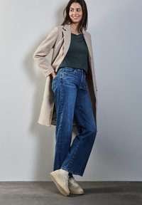 Vaalea beige pitkä takki tummanvihreän paidan päällä, yhdistettynä sinisiin leveälahkeisiin farkkuihin ja beigeihin lenkkareihin. Asusteessa on puhtaat linjat ja tekstuurit.