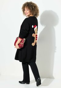 Zwarte gebreide cardigan met een gingerbread man ontwerp op de rug, rode accenten bij de mouwen, gecombineerd met zwarte broek en een rode handtas.