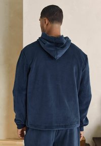 Giacca con cappuccio blu navy realizzata in morbido tessuto testurizzato. Caratterizzata da una vestibilità ampia con polsini a costine e tasca a marsupio. Applicata in uno stile informale.