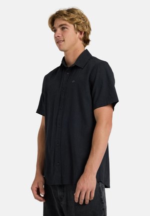 Joven con cabello rubio que lleva una camisa negra de manga corta abotonada y jeans oscuros, de pie y mirando ligeramente hacia la derecha.