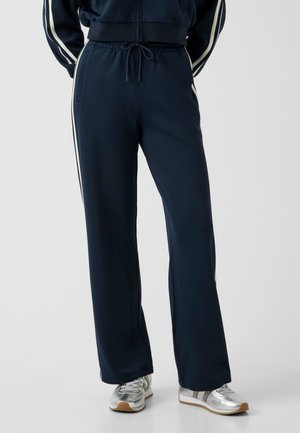 Personne portant un pantalon de survêtement bleu foncé avec des bandes latérales blanches et des baskets argentées, debout sur un fond uni et clair.