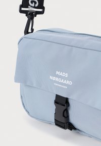 Jasnoniebieska torba na ramię wykonana z trwałego materiału, z klapą składana, czarnymi elementami metalowymi oraz nadrukowanym logo marki na przedzie.