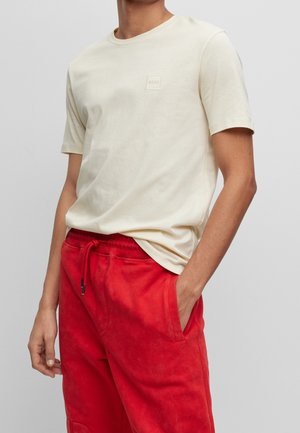 Homme portant un t-shirt crème à manches courtes avec un petit logo et un pantalon rouge vif à cordon, les mains dans les poches.