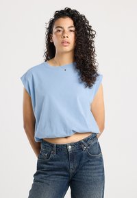 Donna con capelli ricci che indossa una canotta azzurra e jeans blu a vita alta, in piedi con le mani dietro la schiena.