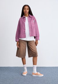 Chaqueta de mezclilla rosa sobre una camiseta blanca, pantalones cortos de pierna ancha marrones y zapatillas naranjas con suelas blancas. Calcetines blancos con franjas moradas.