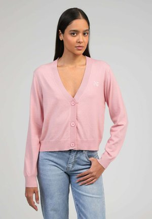 Jeune femme portant un gilet rose boutonné et un jean bleu clair, debout avec une main dans la poche devant un fond uni.