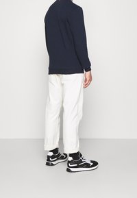 Uomo in felpa blu navy, pantaloni bianchi arrotolati, sneaker in bianco e nero, in piedi di spalle contro uno sfondo bianco.