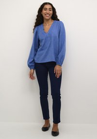 Blouse texturée bleue avec un décolleté en V et des manches bouffantes, associée à un jean skinny bleu marine et des ballerines noires. Design simple, coupe décontractée.