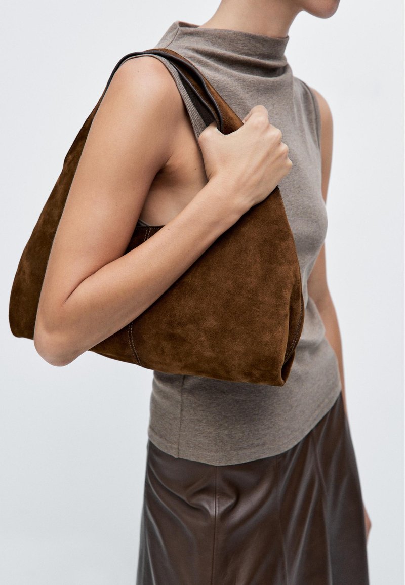 Bruine suede tas met een slouchy ontwerp, featuring afgeronde randen en een zachte textuur, gehouden tegen een minimalistische achtergrond.