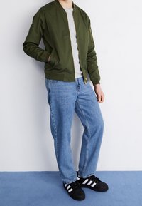 Giubbotto bomber verde oliva con zip, t-shirt grigia, jeans dritti azzurri chiari e sneaker nere con strisce bianche.