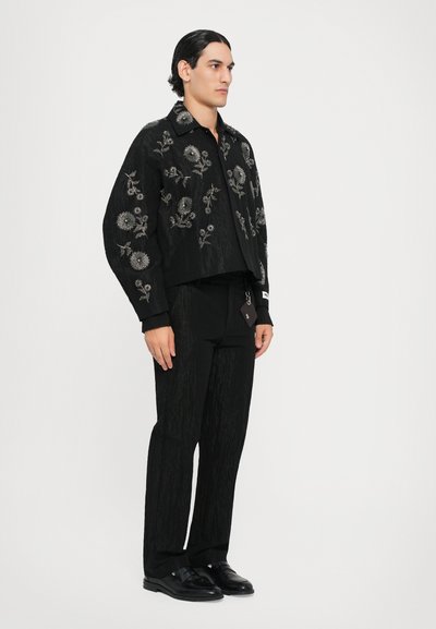 Schwarze bestickte Jacke mit floralen Mustern, kombiniert mit schwarzen Hosen und glänzenden schwarzen Schuhen, verfügt über einen strukturierten Stoff und ein kurzes Design.