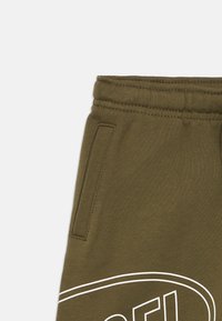 Ελιά πράσινο παντελόνι φόρμας με ελαστική μέση, πλαϊνή τσέπη και λευκό καμπυλωτό σχέδιο κοντά στον μηρό.