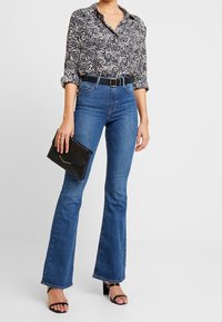 Black and white patterned button-up shirt, blue flared jeans, black belt, and textured black clutch. Styled with black heeled sandals.