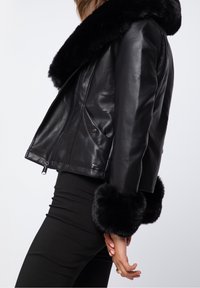 Giubbotto in pelle nera con colletto e polsini in pelliccia sintetica, dal design aderente e chiusura con zip, mostrato di profilo.