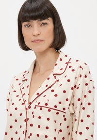 Krémová pyžamová košile s červenými puntíky, kontrastním červeným lemem, klopovým límcem a jednou kapsou na levé straně. Měkká textilie.