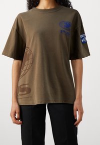 Camiseta de algodón de color verde oliva con mangas cortas. Presenta gráficos en azul y marrón, incluyendo un diseño de un globo y texto, con un parche en la manga.