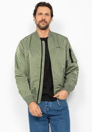 Mand iført olivengrøn bomberjakke over sort skjorte og blå jeans, stående med den ene hånd i lommen mod en hvid baggrund.