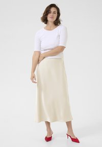 Beige satin midi-nederdel parret med en hvid kortærmet top, komplementeret med røde højhælede sko. Glat tekstur, flydende design.