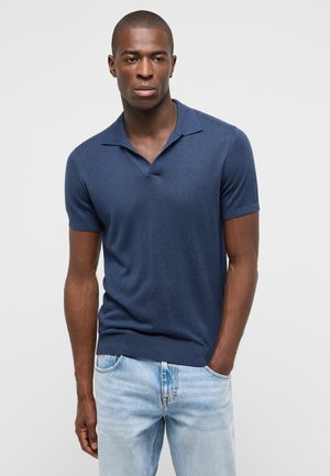 Mann trägt ein marineblaues kurzärmeliges Poloshirt und hellblaue Jeans, steht mit einer Hand in der Tasche vor einem einfarbigen grauen Hintergrund.