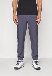 Grijze joggingbroek met elastische boorden, gemaakt van een glad materiaal. Draagt een zwart t-shirt en witte sportschoenen.