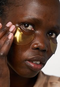 Des patchs oculaires en hydrogel doré appliqués sous les yeux d'une personne à la peau foncée. Texture lisse et brillante sur les patchs ; peau claire et vernis à ongles neutre.