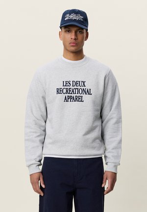 Jeune homme portant un sweat gris avec le texte "Les Deux Recreational Apparel" et une casquette bleu marine, debout devant un fond uni.