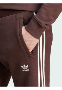 Pantaloni della tuta marroni con dettaglio a tre strisce bianche. Presentano una tasca laterale e un logo Adidas ricamato vicino all'orlo. Texture morbida.