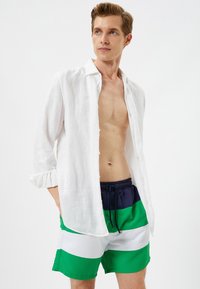 Camisa de lino blanco desabrochada sobre bañadores a rayas en azul y verde. Los pantalones cortos presentan amplias rayas horizontales en verde, blanco y azul marino.