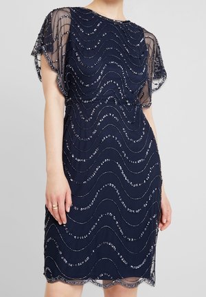 Granatowa sukienka do kolan z przezroczystymi, falującymi rękawami i falistymi wzorami z srebrnych cekinów na tkaninie.
