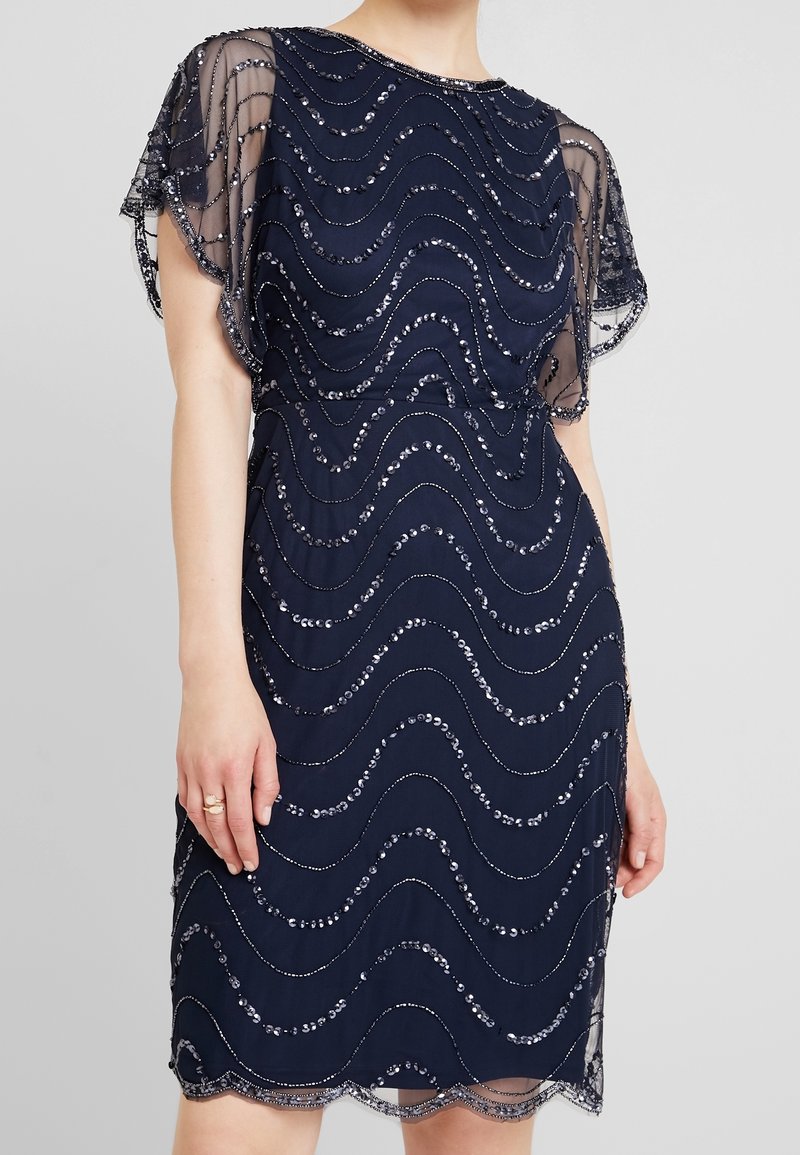 Robe bleu marine longueur genou avec manches volantées en tissu transparent et motifs ondulés de paillettes argentées sur le tissu.