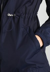 Giacca a vento blu navy con polsini elastici, vita regolabile con corda e tasche laterali. Realizzata in tessuto liscio e leggero. Dettagli delle cuciture visibili.