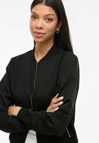 Junge Frau mit langen, dunklen Haaren, die eine schwarze Zip-Jacke und goldene Ohrreifen trägt, steht mit verschränkten Armen vor einem weißen Hintergrund.