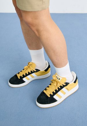 Piernas con pantalones cortos beige, calcetines blancos hasta el tobillo y zapatillas Adidas negras, amarillas y blancas, de pie sobre un suelo azul claro.