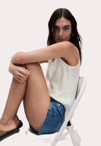 Vit ärmlös stickad topp med texturerad tyg, paired med ljusblå denimshorts med fransade kanter. Ämnet sitter på en vit stol.