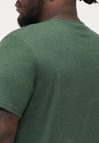 Dunkelgrünes T-Shirt mit kurzen Ärmeln aus strukturierter Stoff, Rundhalsausschnitt und lockerer Passform, mit sichtbaren Nähten.