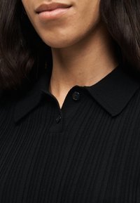 Κοντινό πλάνο του λαιμού και του κάτω μέρους του προσώπου ενός ατόμου που φοράει μαύρο πουλόβερ με ριμπ ύφανση, γιακά και δύο μαύρα κουμπιά.