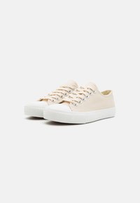 Beige vászon sneaker fehér gumitalppal és orr részen, hat fűzőlyukkal és lapos cipőfűzőkkel. Texturált felsőrész és minimalista dizájn.