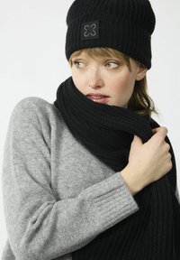 Zwarte gebreide beanie en bijpassende sjaal met een geribde structuur; de trui is lichtgrijs, met geribde boorden en een ontspannen pasvorm.