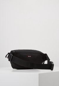 Černá látková crossbody taška se zipovým uzávěrem, nastavitelným popruhem a malým logem v červené, bílé a modré barvě na přední straně. Hladký povrch.