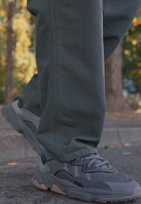 Gray athletic shoes with mesh and synthetic materials, featuring a thick sole, curved design, and textured accents, paired with olive green pants.